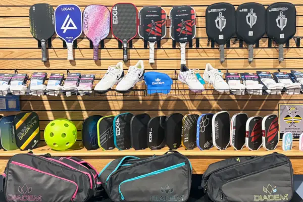 A retail display features various pickleball paddles, shoes, & a blue cap on a wooden wall. Bags & a yellow ball displayed.
